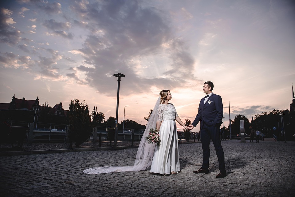Pareja de novios caminando de la mano al atardecer en una sesión de fotos de boda al aire libre, con vestido blanco y ramo, rodeados de un cielo con nubes y luz suave.
